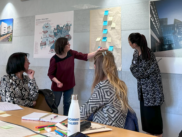 4 personnes sont visibles sur la photo : 2 sont assises à une table et regardent les 2 autres présentant un document sur lequel sont collés plusieurs post-it.