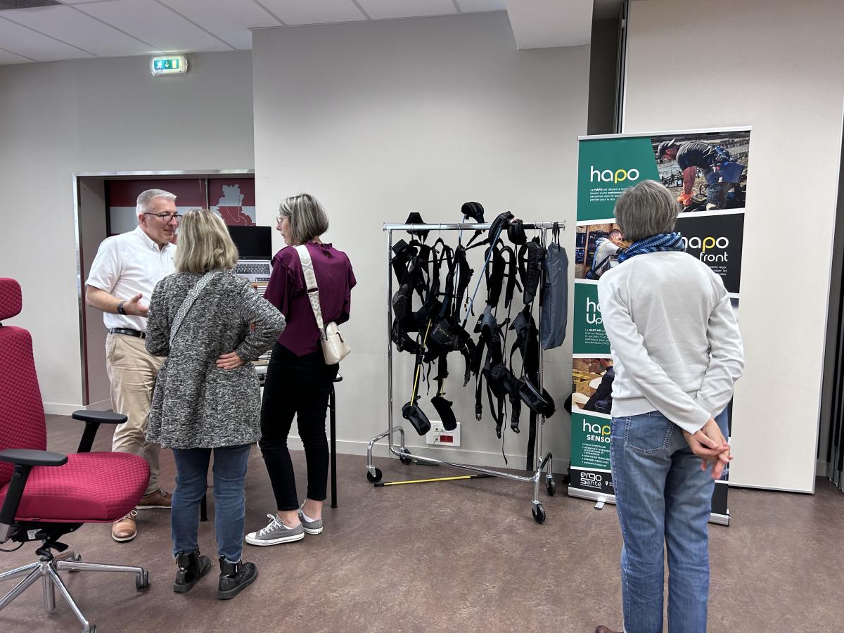 Des exosquelettes sont accrochés sur un portant et plusieurs personnes discutent autour.